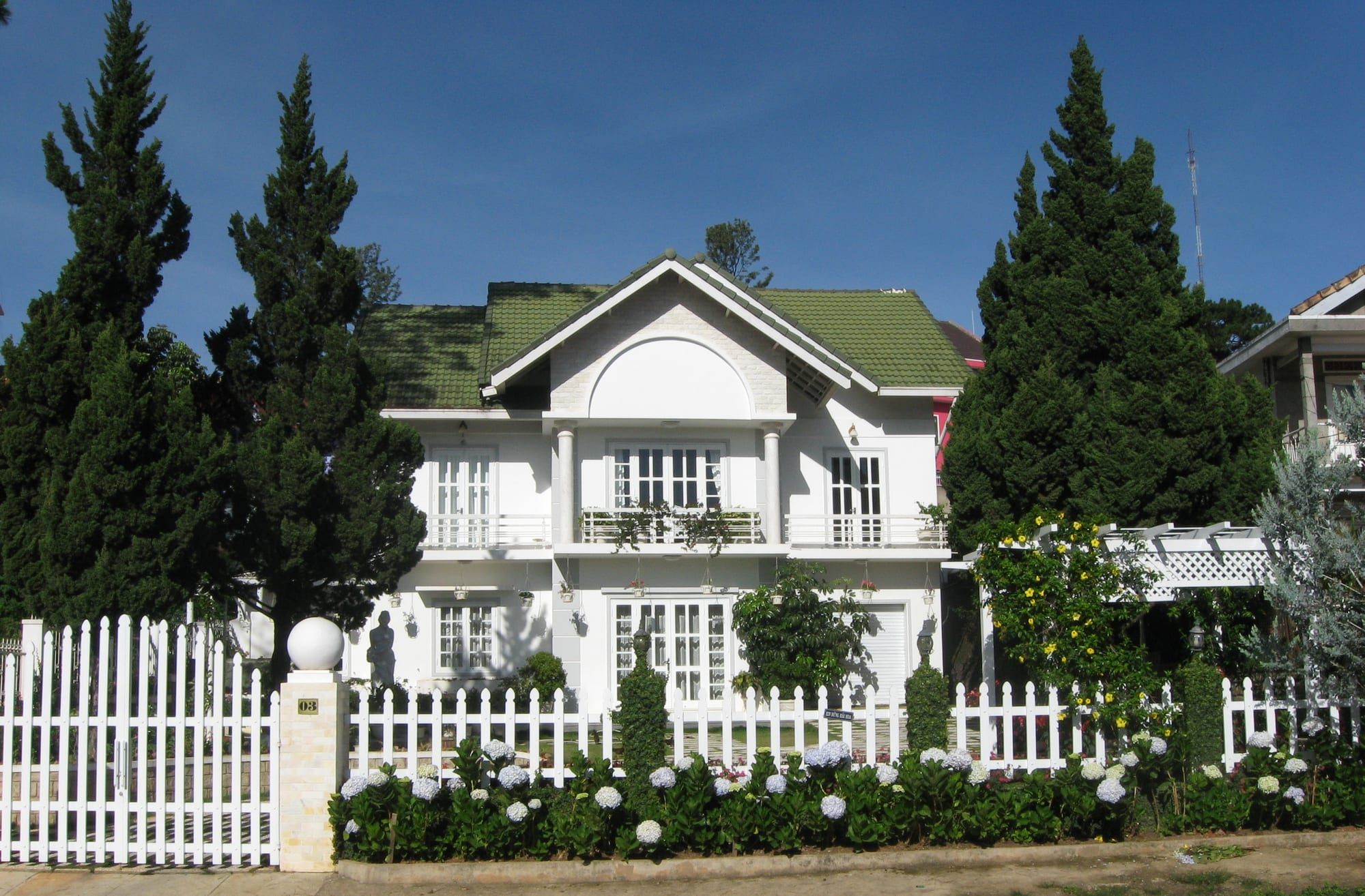 A white two-storey villa with a green tiled roof stands behind a white picket fence, a gate and a bed of hydrangeas, framed by tall cypress trees.