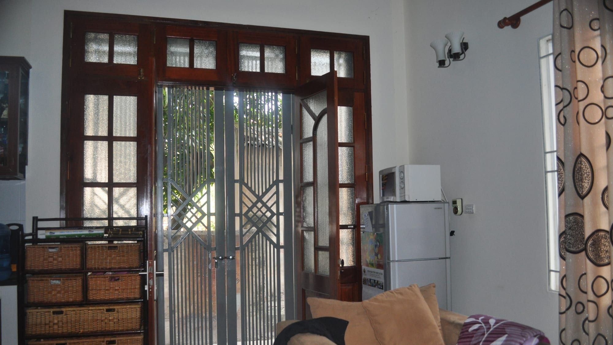A sofa sits in front of tall dark-wood glazed double doors with an iron grille, next to a wicker chest of drawers, a small fridge and a microwave.