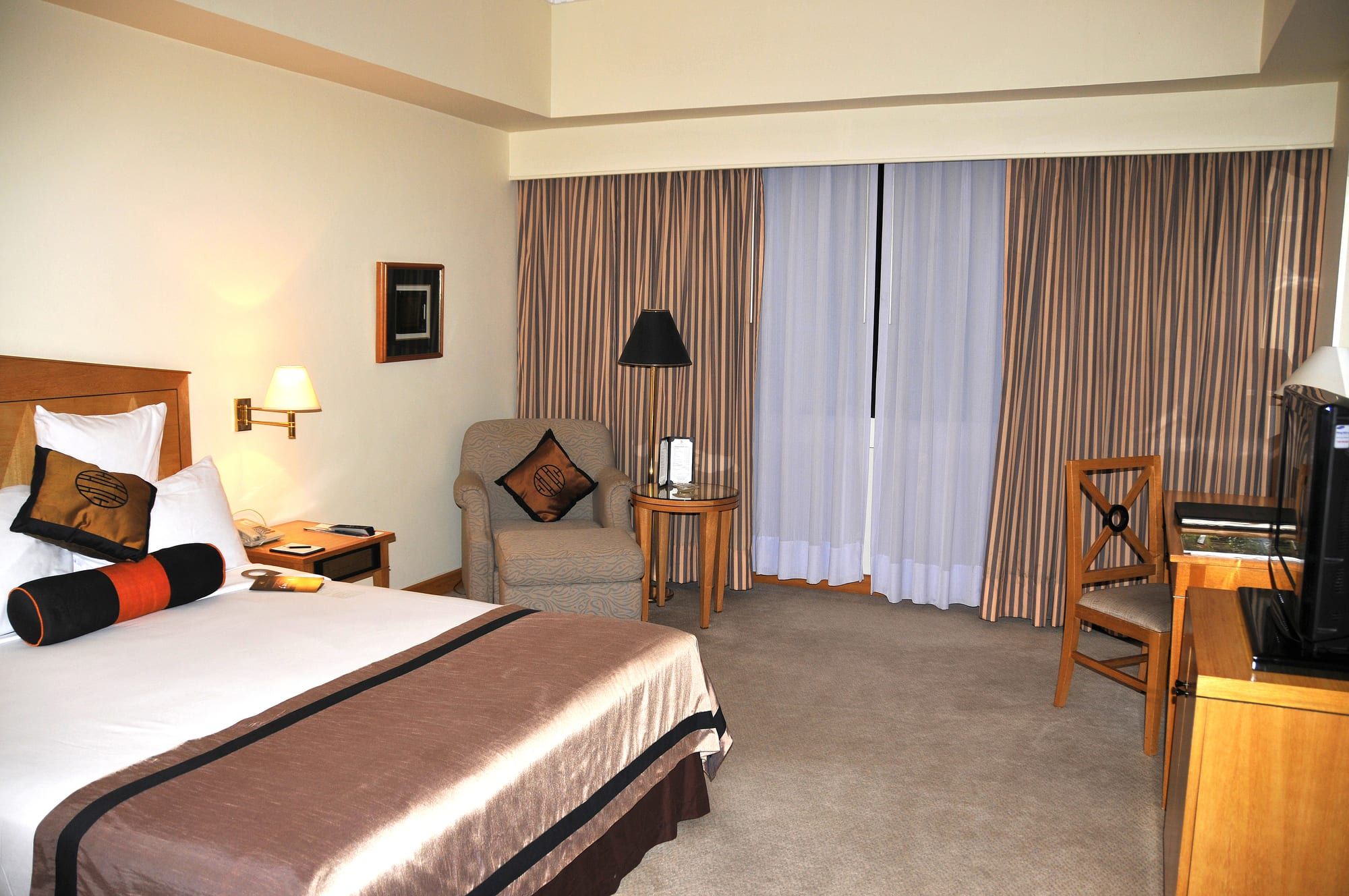 A made double bed with a brown runner and dark cushions, a wall reading lamp, an armchair beside sheer curtains, and a wooden desk with a chair.