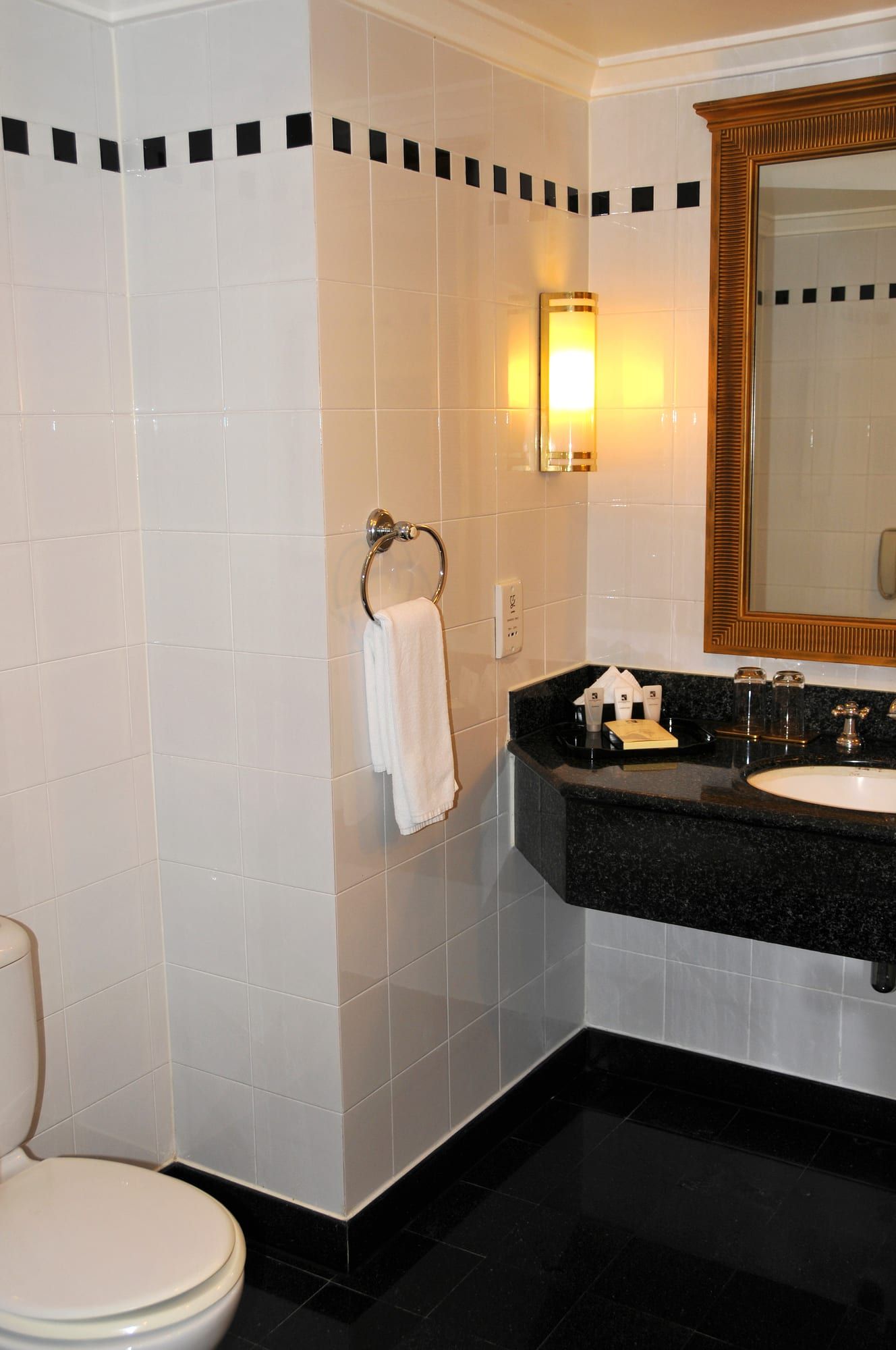 A white-tiled bathroom with a black stone vanity and round basin, a framed mirror, a wall lamp, a hanging towel and a toilet.