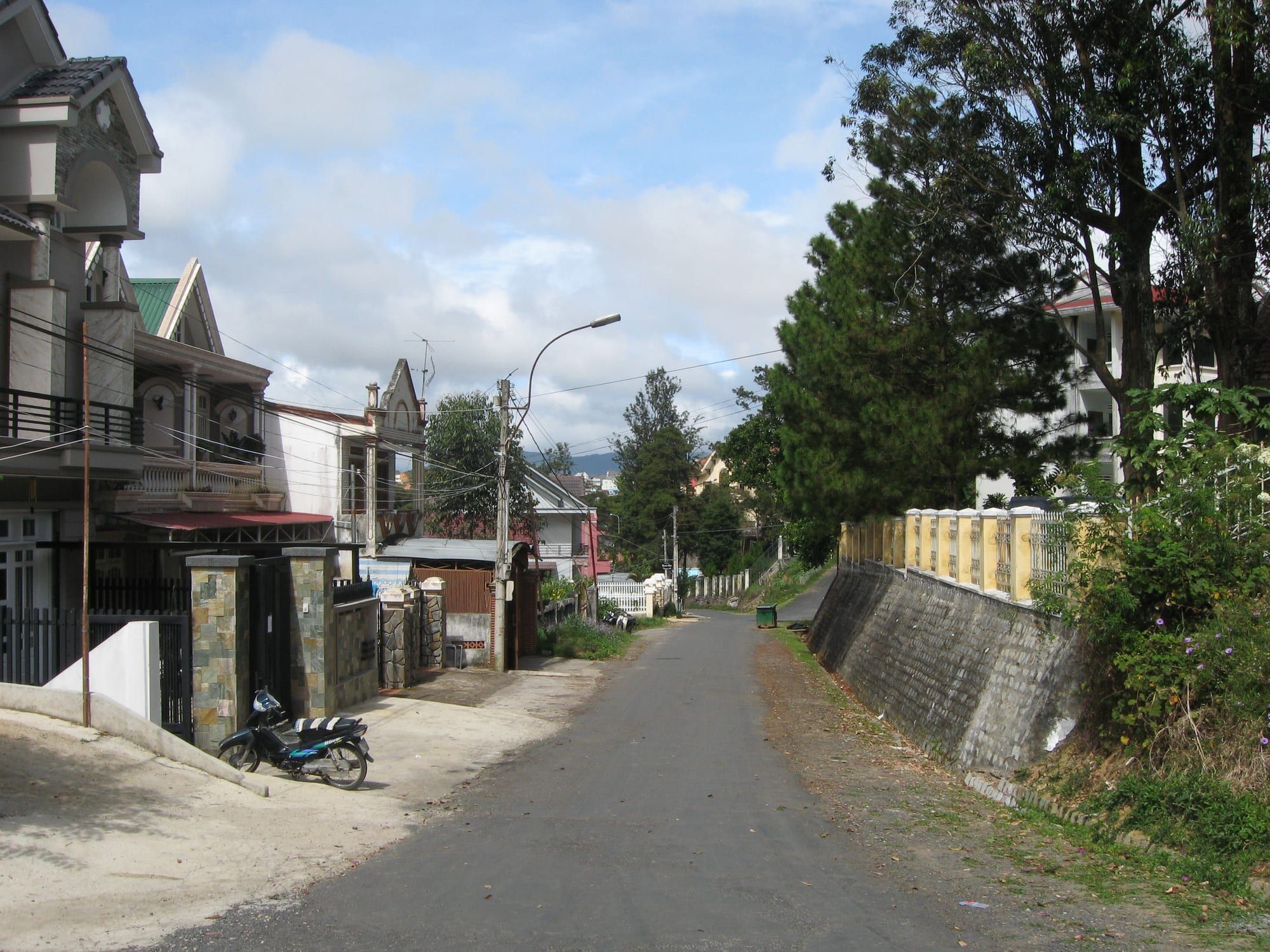 A quiet residential lane runs uphill past gated two-storey houses, a parked motorbike and a stone retaining wall under pine trees.